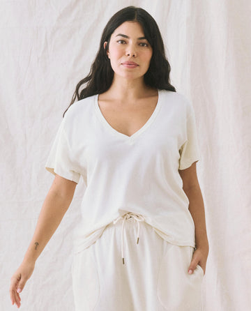 A woman with long dark hair, wearing The V-Neck Tee in Washed White by THE GREAT. SP22 APRILCAP and matching drawstring pants, stands in front of a neutral background. The boxy fit of her outfit adds a touch of effortless style.