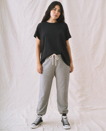 A person stands against a plain background wearing The Boxy Crew in Solid -- Almost Black by THE GREAT. CORE KNITS, gray sweatpants with a drawstring, and black-and-white sneakers.