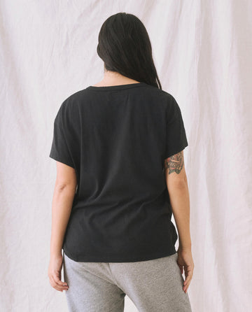 A woman with long dark hair, dressed in The Boxy Crew. Solid -- Almost Black T-shirt from THE GREAT. CORE KNITS and grey sweatpants, stands with her back to the camera against a light-colored fabric backdrop.