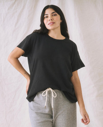 A person wearing The Boxy Crew in Almost Black by THE GREAT. CORE KNITS and gray sweatpants stands against a beige background.
