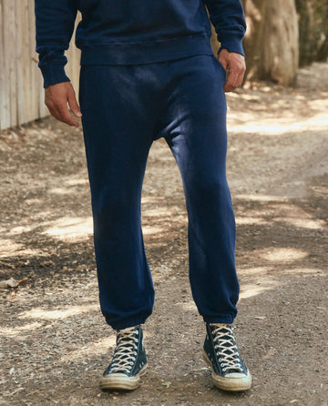 Person wearing vintage athleticwear, including The Men's Stadium Sweatpant in True Navy by THE GREAT. HOL 22 MEN and dark high-top sneakers, standing on a dirt path outdoors.