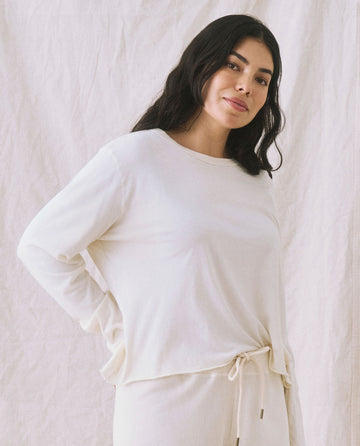 A woman with long dark hair and slim arms, wearing The Long Sleeve Crop Tee in Washed White by THE GREAT. CORE KNITS paired with beige pants, stands in front of a light-colored backdrop.
