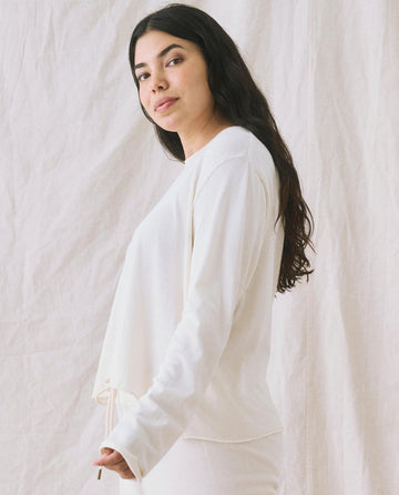 A woman with long dark hair stands against a plain, light-colored fabric backdrop, wearing The Long Sleeve Crop Tee in Washed White by THE GREAT. CORE KNITS.