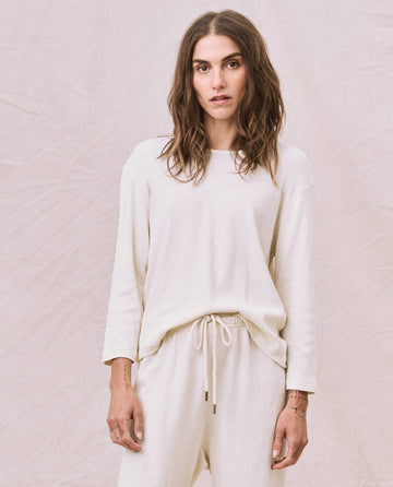 Against a plain background, a person with long hair is wearing THE GREAT. HOL 22 D1's The Long Sleeve Recycled Slim Crew in Natural, crafted from a supersoft and lightweight recycled cotton blend, paired with matching pants.
