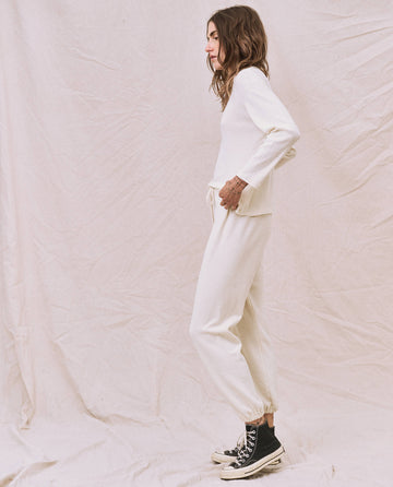 A woman stands sideways, wearing a natural-colored The Long Sleeve Recycled Slim Crew top from THE GREAT. HOL 22 D1, white pants, and black high-top sneakers against a wrinkled white background. The outfit's supersoft and lightweight feel is attributed to the recycled cotton blend fabric.