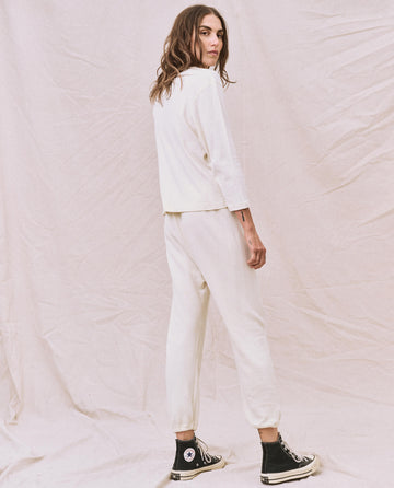 A person with long hair stands sideways wearing The Long Sleeve Recycled Slim Crew in Natural by THE GREAT. HOL 22 D1, paired with all-white casual clothing made from a recycled cotton blend and black high-top sneakers, in front of a neutral backdrop.