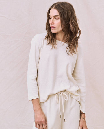 A person with long hair is wearing a light-colored "The Long Sleeve Recycled Slim Crew" top from THE GREAT. HOL 22 D1 and drawstring pants, standing against a neutral background.