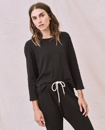 A woman with long hair, dressed in The Long Sleeve Recycled Slim Crew in Night by THE GREAT. HOL 22 D1, pairs it with black pants featuring a white drawstring. She stands against a light-colored fabric backdrop. Crafted locally in L.A., the ensemble boasts supersoft and lightweight comfort.