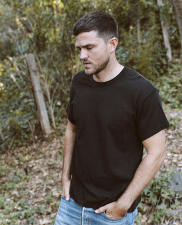 A man in The Men's Boxy Crew by THE GREAT. MAN, almost black, and blue jeans stands outdoors with his hands in his pockets, looking down, surrounded by greenery and trees.