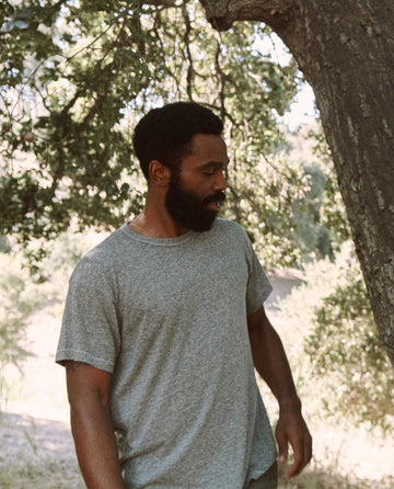 A bearded man wearing The Men's Boxy Crew in heather grey by THE GREAT. MAN stands next to a tree outdoors.
