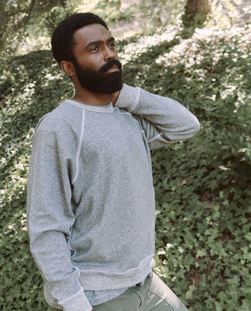 A man with a beard and curly hair stands outdoors in front of greenery, wearing The Men's College Sweatshirt in Varsity Grey from THE GREAT. MAN and looking to the side.