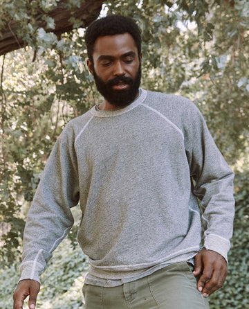 A bearded man stands outdoors amid lush greenery, clad in the varsity grey Men's College Sweatshirt by THE GREAT. MAN.