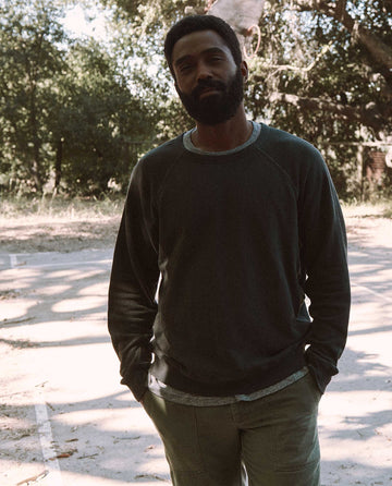 A man stands outdoors on a sunlit, tree-lined path, wearing THE GREAT. MAN's washed black, vintage Men's College Sweatshirt and light-colored pants. Hands in his pockets, he looks directly at the camera.