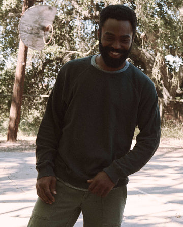 A person with a beard stands outdoors in a wooded area, wearing The Men's College Sweatshirt by THE GREAT. MAN in washed black and light pants. They are looking down and smiling.