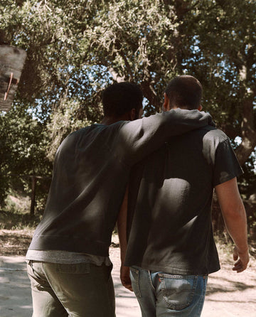 Two people walk closely together on a sunny day, with one person's arm around the other's shoulder, both wearing The Men's College Sweatshirt in washed black by THE GREAT. MAN. Trees and greenery are visible in the background.