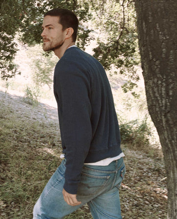 A man with short hair and a beard, dressed in THE GREAT. MAN. Men's College Sweatshirt in washed navy and a pair of jeans, strolls near a tree in a wooded area.