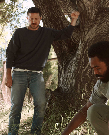 Two men are outdoors, with one standing and the other crouching near an old tree, both focused on the ground in front of them. The man standing wears The Men's College Sweatshirt in Washed Navy from THE GREAT. MAN., adding a nostalgic touch to their curious exploration.