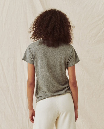 A person with curly hair, wearing The Slim Tee. Solid -- Heather Grey from THE GREAT. CORE KNITS and white pants, stands facing a neutral-colored, textured fabric backdrop.