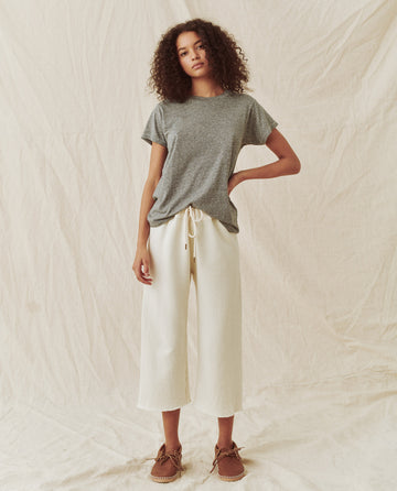 A person with curly hair stands against a plain fabric background, wearing The Slim Tee in Heather Grey by THE GREAT. CORE KNITS, white wide-leg pants, and brown shoes.