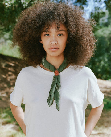 A person with curly hair stands outdoors wearing a white shirt and The Bandana in Army Green, made of ultra-soft hand 100% cotton from THE GREAT. SP24 D2. Trees are visible in the background.
