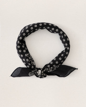 A small "Almost Black" bandana from THE GREAT. SP24 D2, adorned with white floral and paisley patterns, folded and tied in a simple knot, reminiscent of vintage handkerchiefs. Crafted for an ultra-soft feel with 100% cotton.