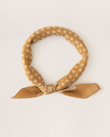 A small, vintage-inspired handkerchief from THE GREAT. SP24 D2, crafted from 100% cotton and featuring a marigold bandana adorned with delicate white floral patterns, neatly tied in a loop with its ends hanging down, displayed on a plain white background.