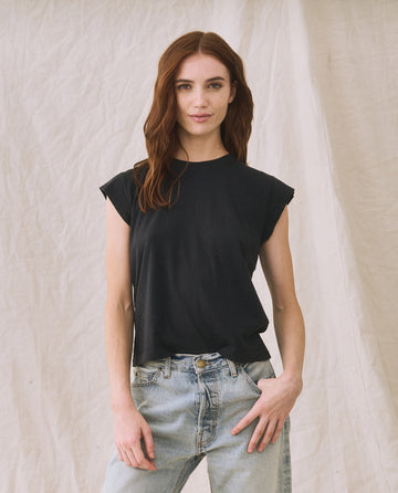 A person with long red hair wearing The Peak Shoulder Tee in Almost Black by THE GREAT. FALL 22 KNITS and high-waisted denim stands against a beige background.