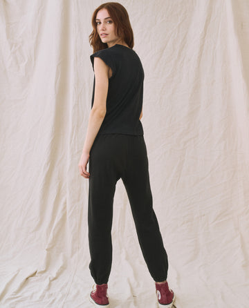 A woman stands in front of a beige backdrop, facing slightly to the side. She is wearing The Peak Shoulder Tee in Almost Black from THE GREAT. FALL 22 KNITS collection, black pants, and red high-top sneakers.