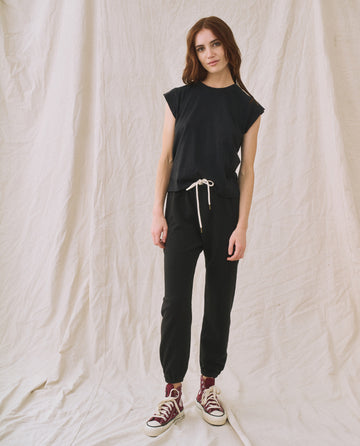 A person stands in front of a beige fabric backdrop, wearing The Peak Shoulder Tee in Almost Black from THE GREAT. FALL 22 KNITS, paired with high-waisted denim and red sneakers.