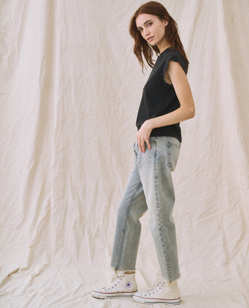 A woman with auburn hair is standing sideways, wearing "The Peak Shoulder Tee" in Almost Black by THE GREAT. FALL 22 KNITS, paired with high-waisted denim jeans and white sneakers. The background features light-colored draped fabric.