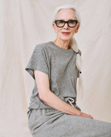 An elderly woman with long white hair in a braid, wearing black-rimmed glasses and The Boxy Crew in Heather Grey from THE GREAT. CORE KNITS, sits against a neutral background, showcasing her timeless style with subtle flattering tweaks.
