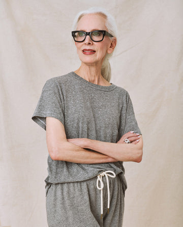 An older woman with white hair, wearing glasses and sporting The Boxy Crew in Heather Grey from THE GREAT. CORE KNITS, stands with her arms crossed in front of a beige background.