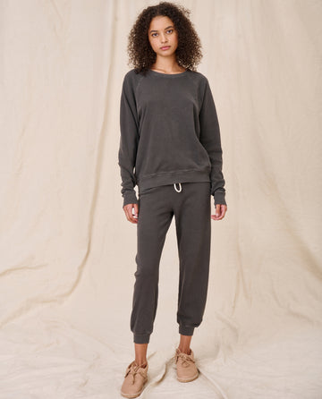 A person with curly hair stands against a neutral background, wearing a matching The College Sweatshirt in Washed Black from THE GREAT. CORE KNITS, along with dark gray joggers and beige shoes.