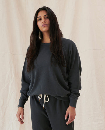 A person with long dark hair wearing "The College Sweatshirt. Solid -- Washed Black" from THE GREAT. CORE KNITS and matching sweatpants stands against a beige fabric backdrop.
