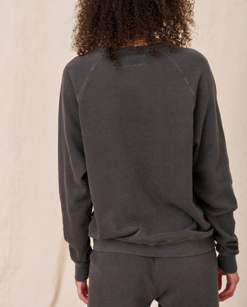 Person with curly hair facing away, wearing THE GREAT. CORE KNITS' The College Sweatshirt in Washed Black and matching pants, standing against a light background.