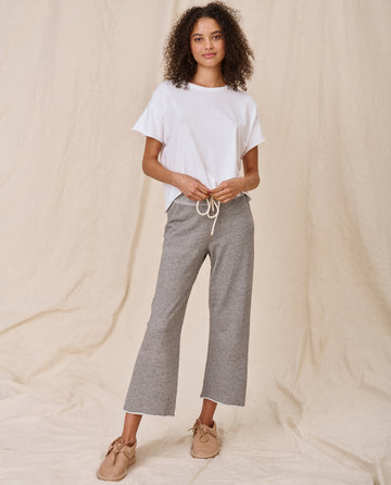 A person with curly hair stands against a beige backdrop, wearing The Crop Tee in True White by THE GREAT. CORE KNITS, gray drawstring pants, and beige shoes.