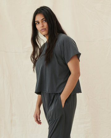 A woman with long dark hair stands against a beige background, wearing The Crop Tee in Washed Black by THE GREAT. CORE KNITS, paired with loose-fitting high-rise jeans made from super soft cotton.
