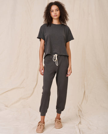 A woman with curly hair is standing against a plain backdrop, wearing The Crop Tee in Washed Black by THE GREAT. CORE KNITS, matching jogger pants, and beige shoes.