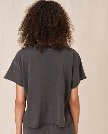 Back view of a person with curly hair wearing a loose-fitting, short-sleeve dark gray 'The Crop Tee' from THE GREAT. CORE KNITS, made from super soft cotton and matching pants, standing against a beige background.