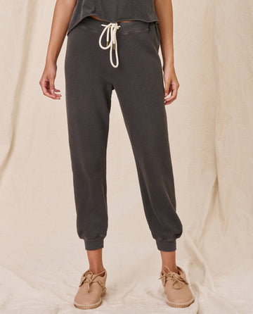 A person wearing The Cropped Sweatpant in Washed Black with ribbed cuffs and a drawstring waistband from THE GREAT. CORE KNITS stands against a beige fabric background, paired perfectly with beige shoes.