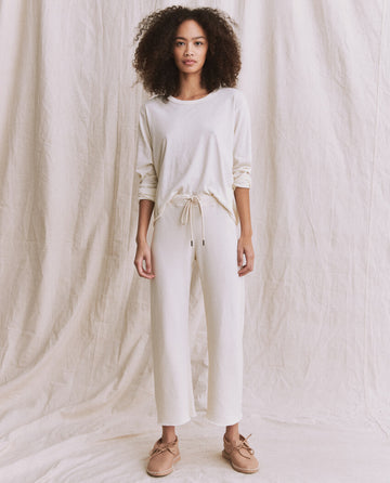 A person with curly hair stands against a light fabric backdrop, donning The Long Sleeve Crop Tee in Washed White by THE GREAT. CORE KNITS, highlighting their long slim arms and matching pants, styled with beige loafers.