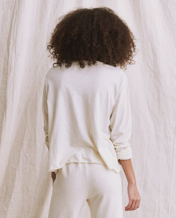 Person with curly hair, seen from behind, wearing The Long Sleeve Crop Tee in Washed White by THE GREAT. CORE KNITS and light-colored pants, standing against a plain fabric backdrop.