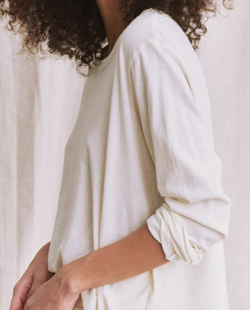 A person with curly hair and long slim arms wearing The Long Sleeve Crop Tee in washed white from THE GREAT. CORE KNITS stands against a light background.