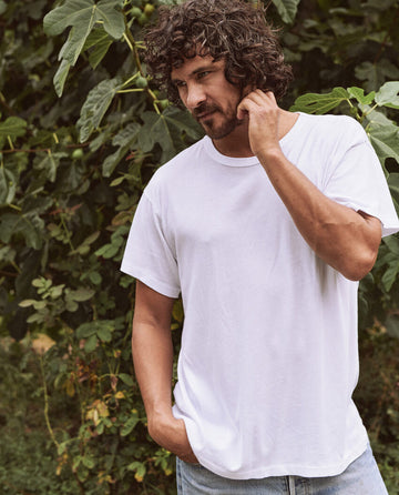 A man with curly hair wearing The Men's Boxy Crew in TRUE WHITE from THE GREAT. MAN stands outdoors, with one hand in his pocket and the other touching his ear, surrounded by green foliage.