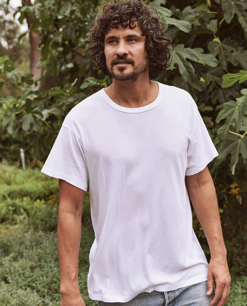 A man with curly hair and a beard is standing outdoors near green foliage, effortlessly cool in his classic staple, The Men's Boxy Crew in True White from THE GREAT. MAN.