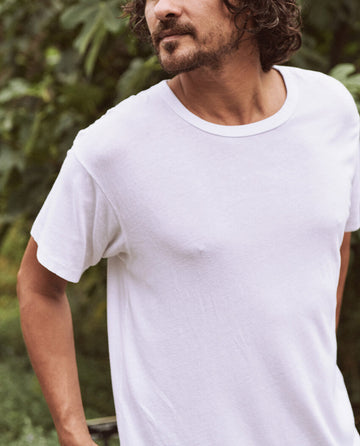 A person with curly hair and a beard is standing outside, effortlessly cool in The Men's Boxy Crew tee by THE GREAT. MAN in true white. The background features lush green foliage.