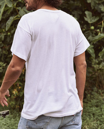 A person with medium-length hair stands outdoors with their back to the camera, wearing a timeless piece from THE GREAT. MAN—The Men's Boxy Crew in TRUE WHITE—paired with blue jeans. Green foliage is visible in the background, creating an effortlessly cool scene.