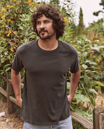 A man with curly hair and a beard is standing outdoors in front of plants, wearing The Men's Boxy Crew in Washed Black by THE GREAT. MAN and jeans, with his hands behind his back.