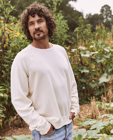 A man with curly hair and a beard stands outdoors in a garden. He is wearing The Men's College Sweatshirt in washed white by THE GREAT. MAN, along with blue jeans, with one hand in his pocket. Trees and plants are in the background.