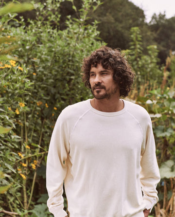 A person with curly hair and a beard is standing outdoors in front of foliage, wearing THE GREAT. MAN's The Men's College Sweatshirt in washed white.